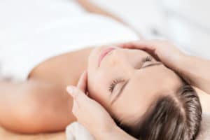 woman lying down on table wrapped in towel as professional runs hands over her head with solid white background